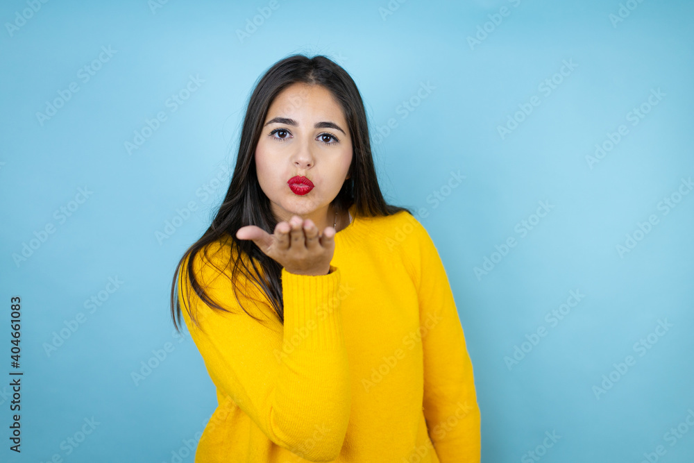 Young beautiful woman wearing yellow sweater over isolated blue background looking at the camera blowing a kiss with hand on air being lovely and sexy. Love expression. Young beautiful woman wearing yellow sweater over isolated blue background looking at the camera blowing a kiss with hand on air being lovely and sexy. Love expression.
