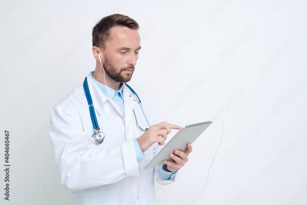 Young serious doctor in earphones holding tablet while taking online patients Young serious doctor in earphones holding tablet while taking online patients