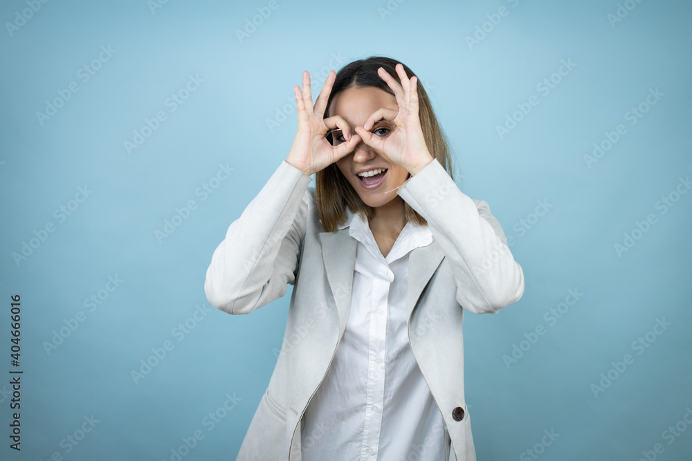 Young business woman over isolated blue background doing ok gesture shocked with smiling face, eye looking through fingers Young business woman over isolated blue background doing ok gesture shocked with smiling face, eye looking through fingers