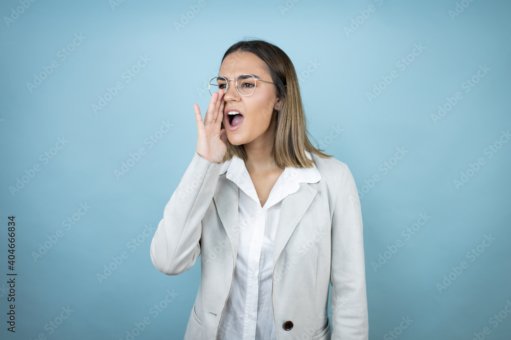 Young business woman over isolated blue background shouting and screaming loud to side with hand on mouth Young business woman over isolated blue background shouting and screaming loud to side with hand on mouth