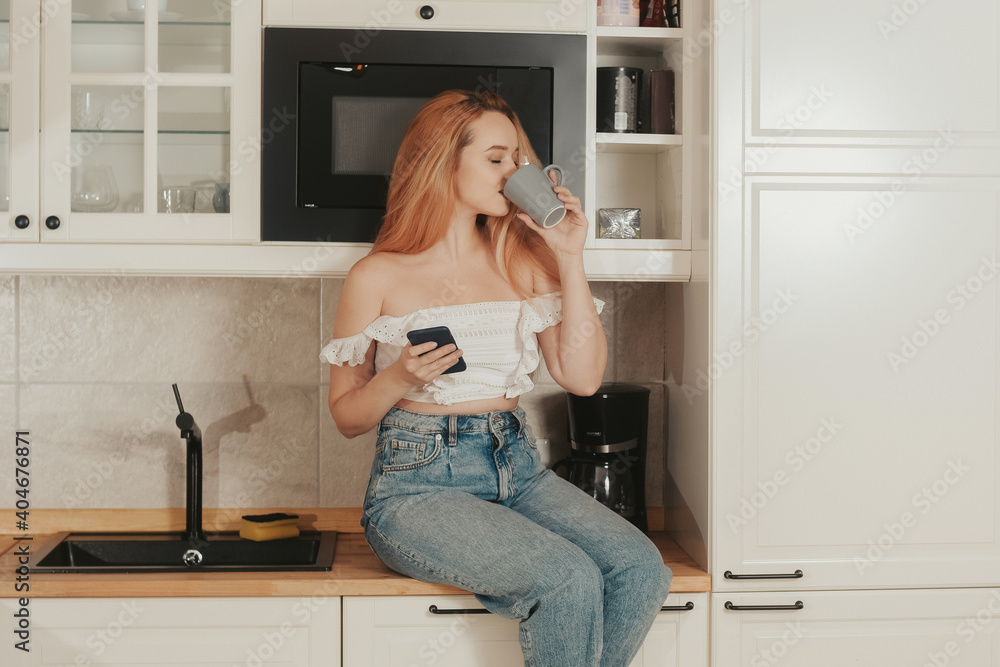 The woman is sitting at home in the kitchen. A girl with a good figure is drinking tea and looking at the phone. Food delivery, mobile shops, online shopping. Drinks tea or coffee The woman is sitting at home in the kitchen. A girl with a good figure is drinking tea and looking at the phone. Food delivery, mobile shops, online shopping. Drinks tea or coffee