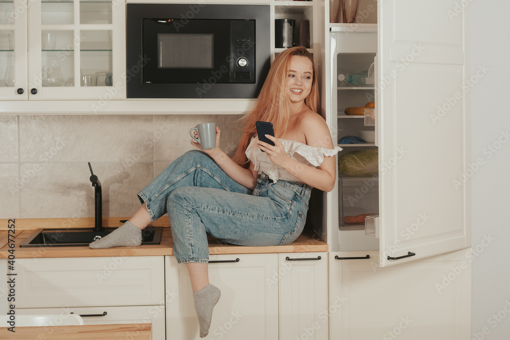 A woman is sitting at home in the kitchen at the will of the refrigerator. A girl with a good figure holds a mobile phone in her hands. Food delivery, mobile shops, online shopping. A woman is sitting at home in the kitchen at the will of the refrigerator. A girl with a good figure holds a mobile phone in her hands. Food delivery, mobile shops, online shopping.
