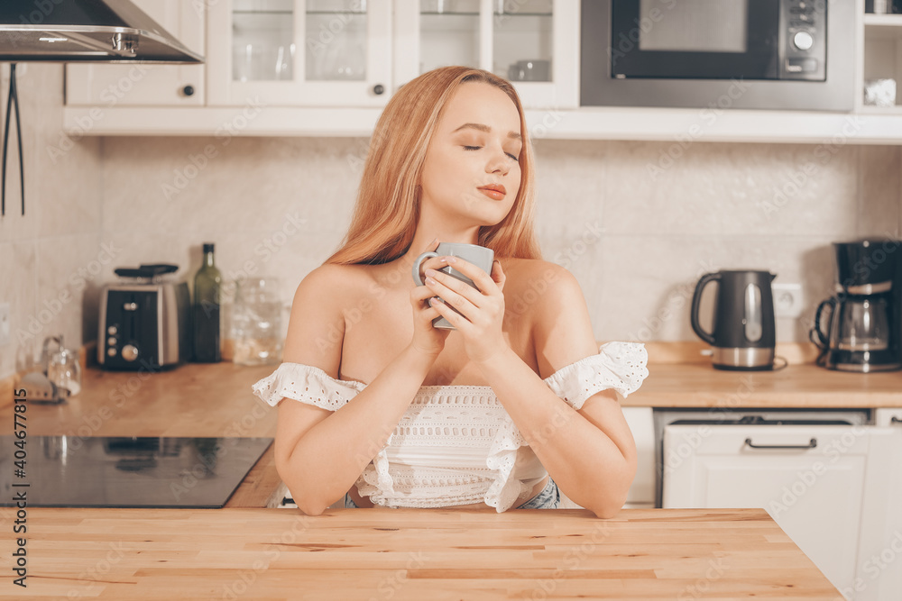 Beautiful woman sits at a table on the background of the kitchen at home. A girl with delight and closed eyes drinks tea or coffee or water in the morning. Beautiful woman sits at a table on the background of the kitchen at home. A girl with delight and closed eyes drinks tea or coffee or water in the morning.