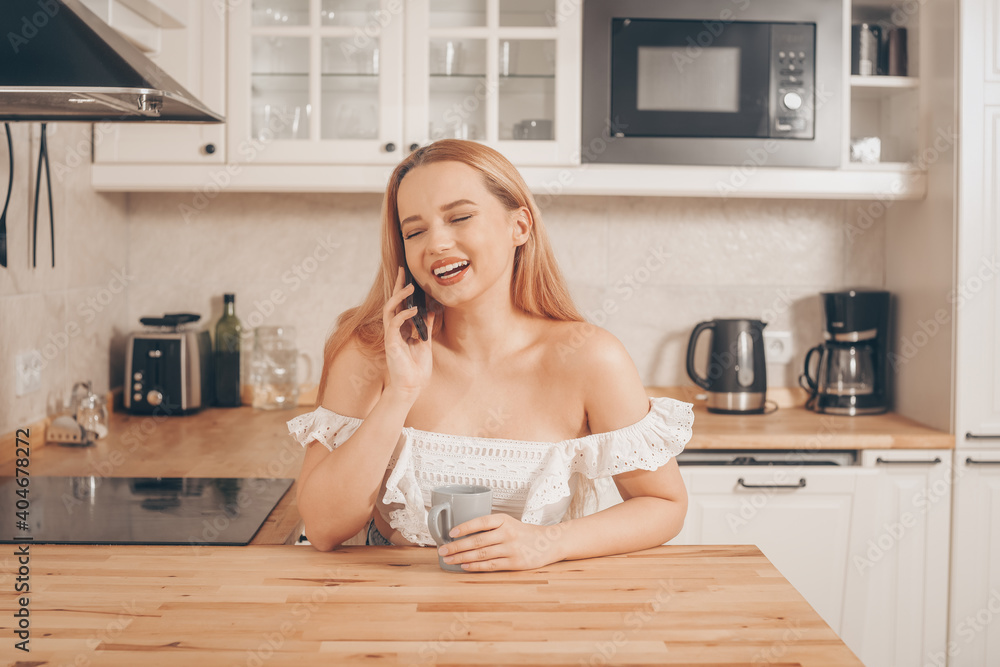 A beautiful girl is sitting in the kitchen drinking tea or coffee and looking at the phone. Online shopping, delivery from food stores. The girl speaks on a smart phone and smiles A beautiful girl is sitting in the kitchen drinking tea or coffee and looking at the phone. Online shopping, delivery from food stores. The girl speaks on a smart phone and smiles