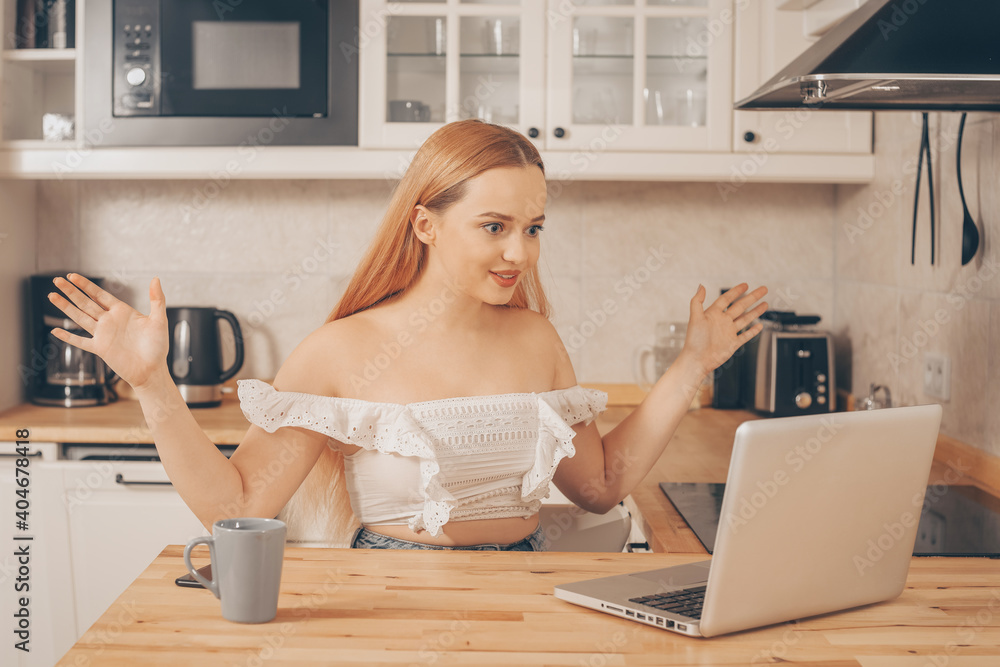 The woman communicates via video, gestures to the webcam, conducts an online lecture. A beautiful girl sits at home in the kitchen and works at a laptop. There is a cup of coffee or tea nearby. The woman communicates via video, gestures to the webcam, conducts an online lecture. A beautiful girl sits at home in the kitchen and works at a laptop. There is a cup of coffee or tea nearby.