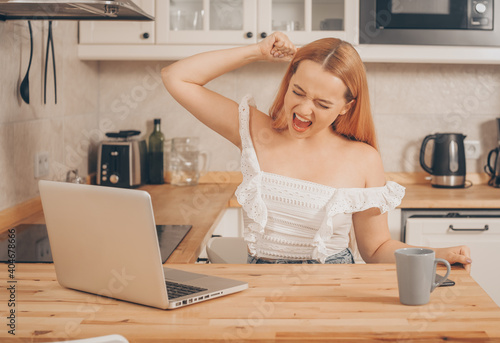 The woman looks into the laptop and rejoices, the girl sits at home against the background of the kitchen and smiles. The blonde won something online, found a job online.