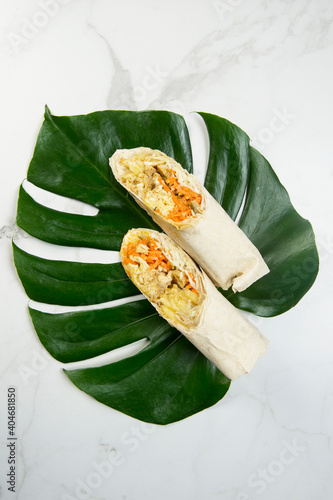 From above of chicken shawarma roll with vegetables (carrot, cucumber, cabbage) cheese wrapped in thin lavash bread. Grilled gyros served on monstera leaf on marble background. Eastern street food
 photo