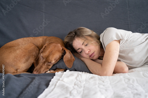 Fotografie Unhappy depressed woman lying with lovely Vizsla dog at home on sofa, thinking pondering, feels apathy and indifference to everything, has mental problems, melancholy attack
