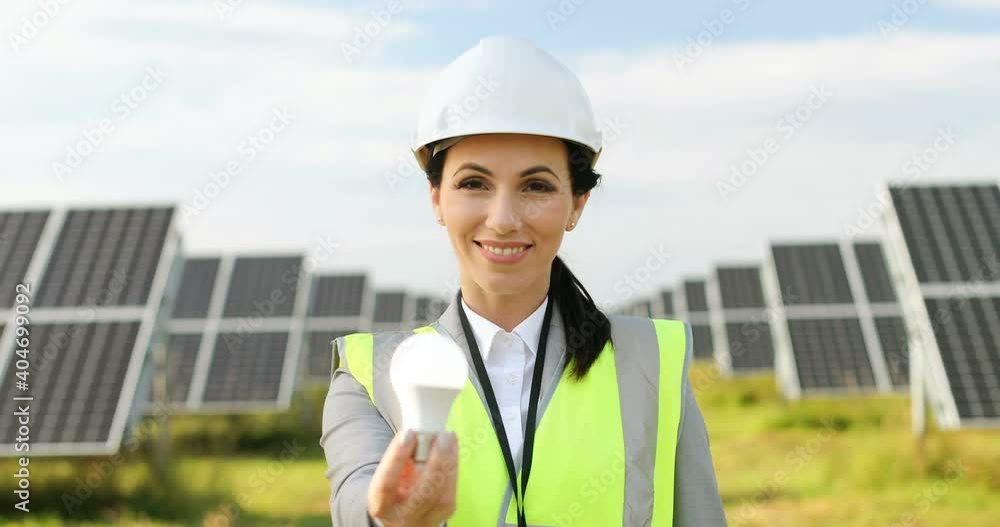 Close up portrait of beautiful female technologist in uniform among field of solar panels. Cheerful woman reaches out holding light bulb. Close up portrait of beautiful female technologist in uniform among field of solar panels. Cheerful woman reaches out holding light bulb.