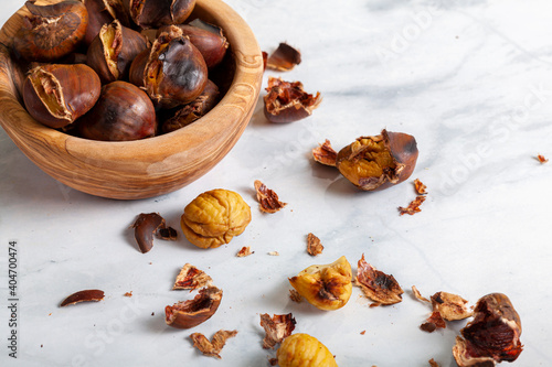 A wooden bowl of homemade oven roasted tsty chestnuts with shells and skins scattered on the marble table top. A traditional winter, autumn snack with a unique flavor. Commonly eaten during christmas