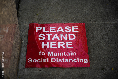 The words "Please Stand Here To Maintain Social Distancing" are placed on the sidewalk outside of a business to help stop the spread of Covid-19.