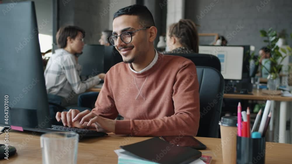 Slow motion of cheerful Arab man using computer typing looking at screen sitting at desk in office while people are working in background. Technology and workplace concept.