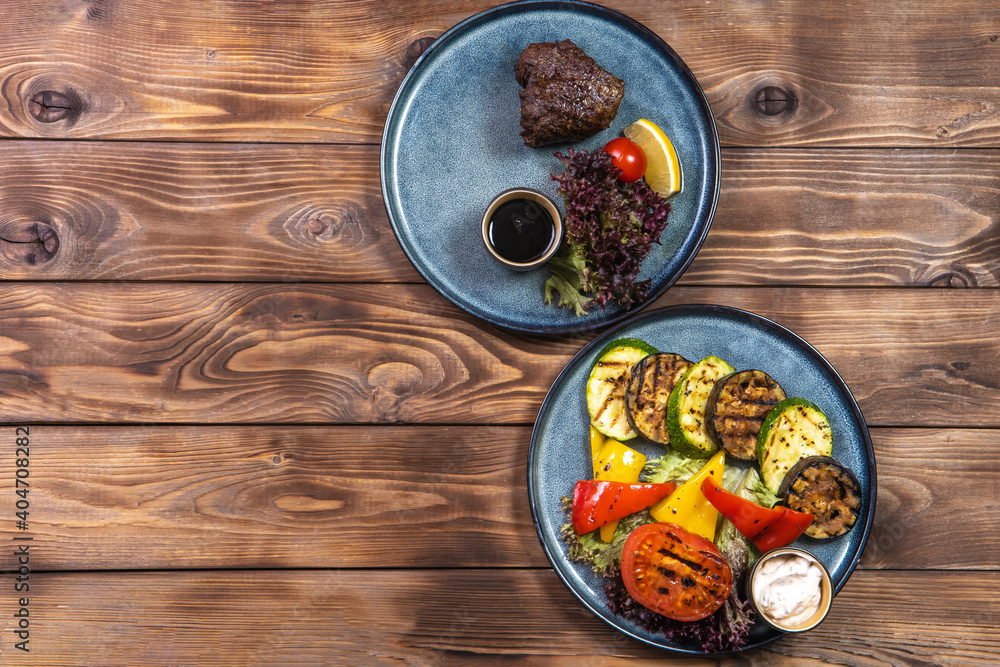 Beef steak with lemon, tomato, lettuce and grilled vegetables on plates on a brown wooden background. Flat layout. Beef steak with lemon, tomato, lettuce and grilled vegetables on plates on a brown wooden background. Flat layout.