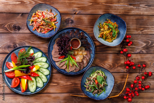 Flat layout of various dishes with sliced vegetables, meat cuts, salads on a brown wooden background.
