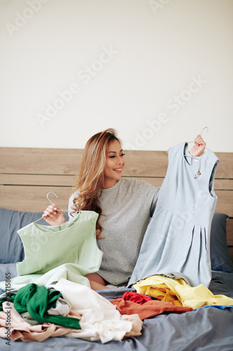 Pretty young woman sitting on bed with many clothes around and deciding what to wear for event