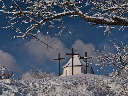 Beautiful view of the top of Kornbühl hill, Swabian Alb, Germany in winter season with snow-covered branches and historic chapel Salmendinger Kapelle and three Christian crosses in front on sunny day. photo