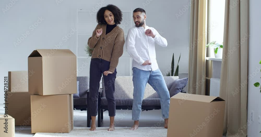 Cardboard boxes stand in living room, young adult couple owners buyers dance together make identical movements with hands standing near parcels, concept of joy and victory, obtaining housing loan Cardboard boxes stand in living room, young adult couple owners buyers dance together make identical movements with hands standing near parcels, concept of joy and victory, obtaining housing loan