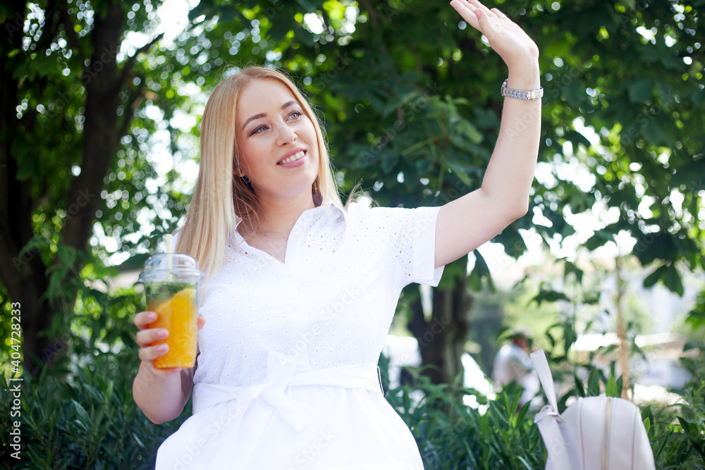 Fototapeta premium Beautiful overweight lady in white dress and holding the bagpack waking in the street of the city and smiling. Plus size girl drink orange cocktail in the park