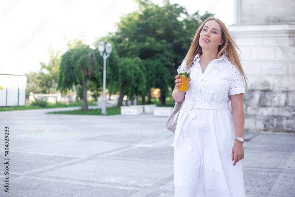 Beautiful overweight lady in white dress and holding the bagpack waking in the street of the city and smiling. Plus size  girl drink orange cocktail in the park