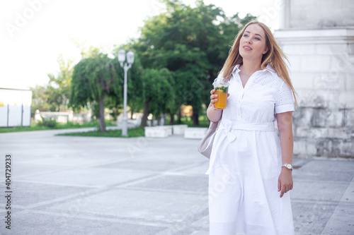 Beautiful overweight lady in white dress and holding the bagpack waking in the street of the city and smiling. Plus size girl drink orange cocktail in the park