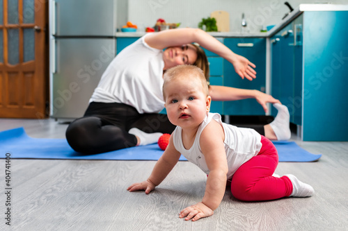 The child crawls on the floor. In the background, in a blur, a young mother is stretched out on a rug in the kitchen. Concept of home sports training with children