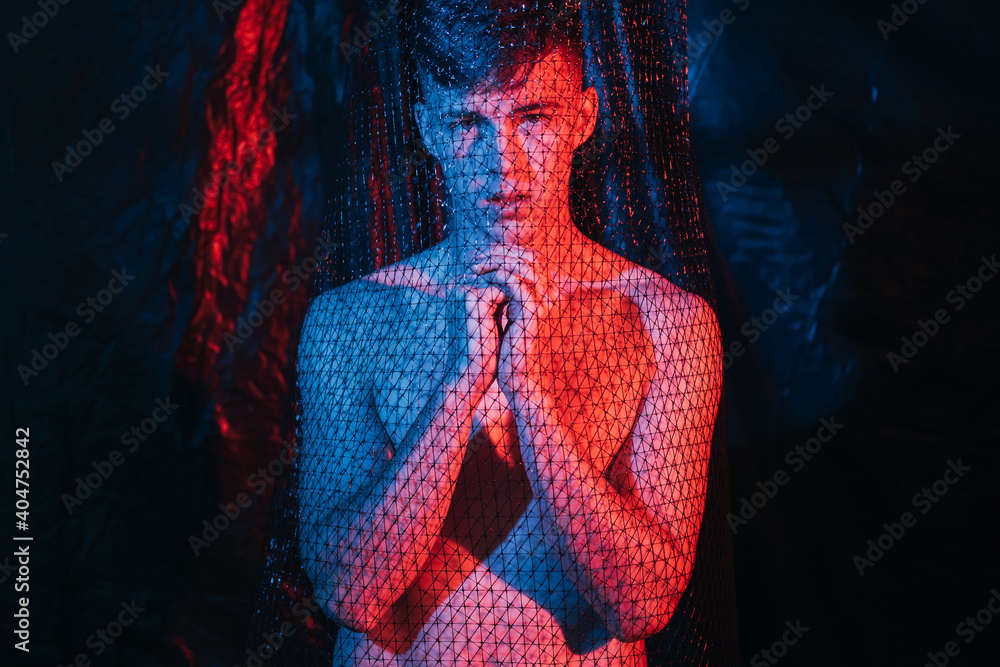 Hopeless man. Human trafficking. Protection security. Disturbed shirtless  male victim praying to God for help forgiveness in mesh trap in red blue  neon light at night on blur dark background. Photos