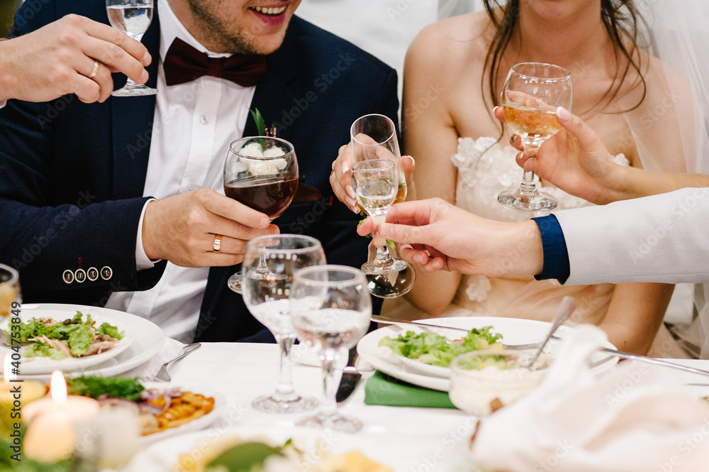 Cheers! Newlyweds with friends drink champagne celebrate at the table. People raise glasses of wine and vodka for toast. Group of men and women celebrate wedding. Cheers! Newlyweds with friends drink champagne celebrate at the table. People raise glasses of wine and vodka for toast. Group of men and women celebrate wedding.