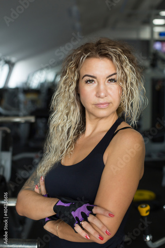 Woman with long blonde curly hair - portrait of a fitness trainer in the gym. Sports form, healthy lifestyle, muscle building, weight loss. Sports wear, gloves without fingers. Beauty and confidence