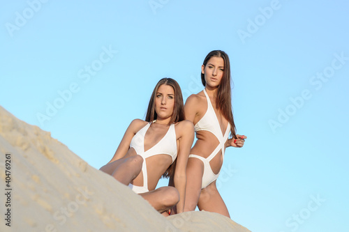 Twins on the beach wearing fashion beachwear. Two perfectly shaped young woman twins sunbathing by the sea.