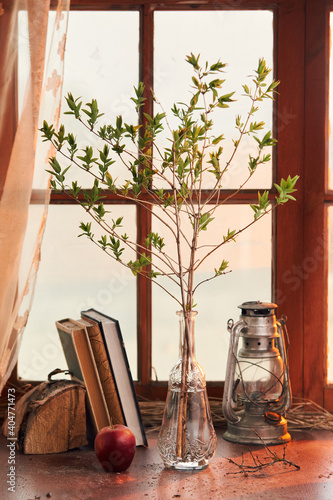 spring twig in a transparent glass vase with a kerosene lamp and books on the background of the window (ID: 404771473)