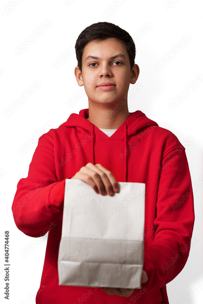 Teenage boy holding white paper bag in his hand isolated on white background. Young caucasian guy wearing red hoodie offering package with content. Vertical orientation.