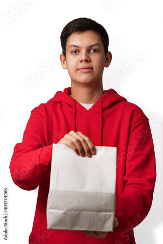 Obraz na płótnie Teenage boy holding white paper bag in his hand isolated on white background. Young caucasian guy wearing red hoodie offering package with content. Vertical orientation. w salonie