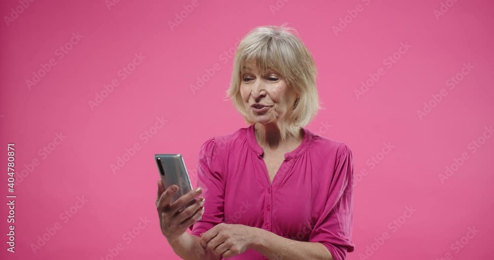 Close up portrait of cheerful happy old Caucasian smiling pretty blonde woman stand in studio talking on video chat on smartphone isolated on pink background videochatting on cellphone and waving hand Close up portrait of cheerful happy old Caucasian smiling pretty blonde woman stand in studio talking on video chat on smartphone isolated on pink background videochatting on cellphone and waving hand
