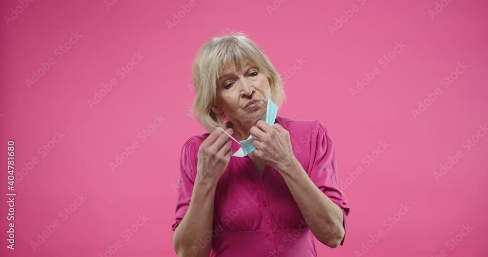 Close up portrait of unhappy Caucasian elderly pensioner blonde woman putting on face medical mask standing posing on pink background isolated in studio alone. Coronavirus quarantine Close up portrait of unhappy Caucasian elderly pensioner blonde woman putting on face medical mask standing posing on pink background isolated in studio alone. Coronavirus quarantine