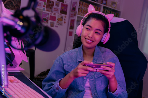 Young playful Asian woman sitting in front of her computer workstation and smiling at the screen with a microphone in foreground. Beautiful asian woman working from her home studio and playing game. photo