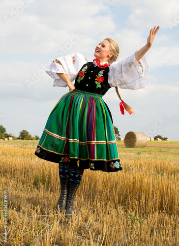 Blonde pretty young smiling girl dressed in Polish national folk dress dancing and spinning around in the field of harvest straw bales photo