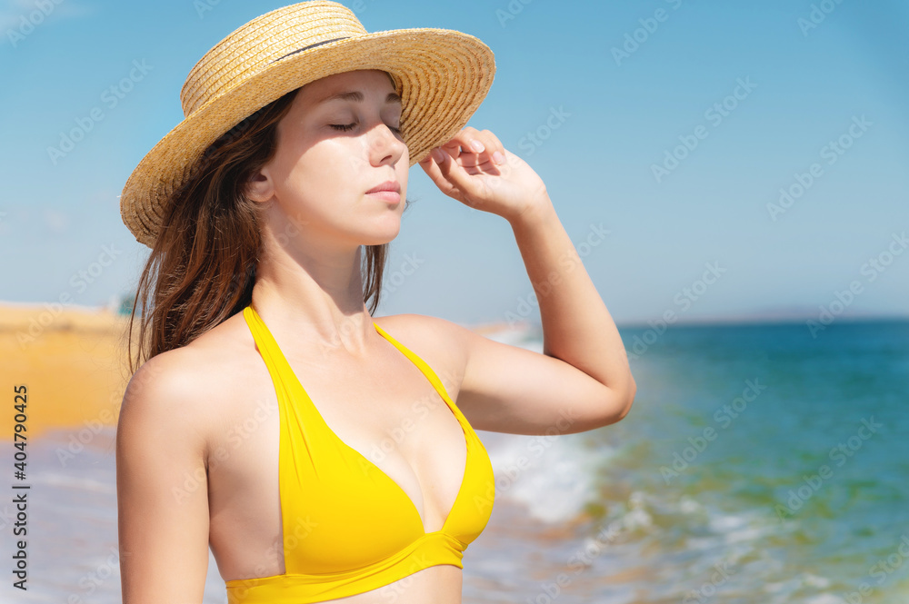 Attractive young woman in a yellow swimsuit and a straw hat. Holding on to the hat against the background of a golden beach and azure sea