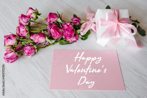 Composition of pink small roses, gift box, postcard on white wooden background. Holiday greeting card with text happy Valentine's day.