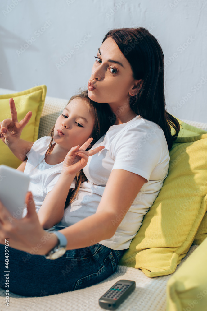 Mother with pouting lips and daughter showing peace gesture taking selfie on couch on blurred foreground