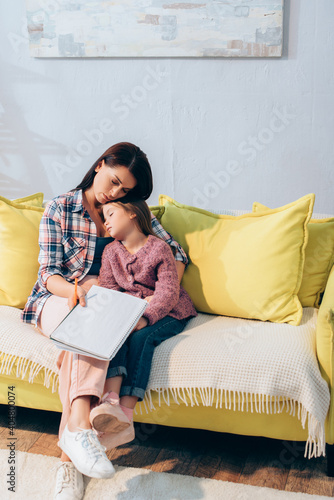 Full length of sad mother with copy book hugging daughter sleeping on couch at home