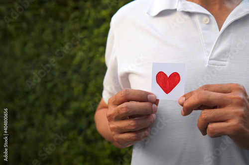 Man’s hand holding hand draw red heart on sliced piece of white paper left side with blurred bush nature out background