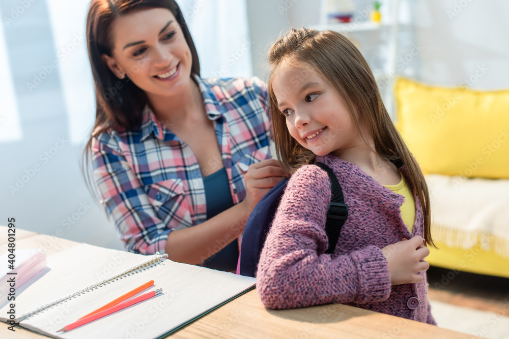 Happy mother putting backpack on daughter near desk at home on blurred background Happy mother putting backpack on daughter near desk at home on blurred background