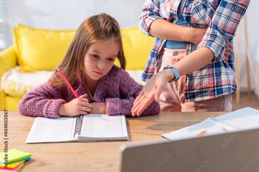 Upset daughter with pencil and notebook near mother pointing with hand near desk on blurred foreground Upset daughter with pencil and notebook near mother pointing with hand near desk on blurred foreground