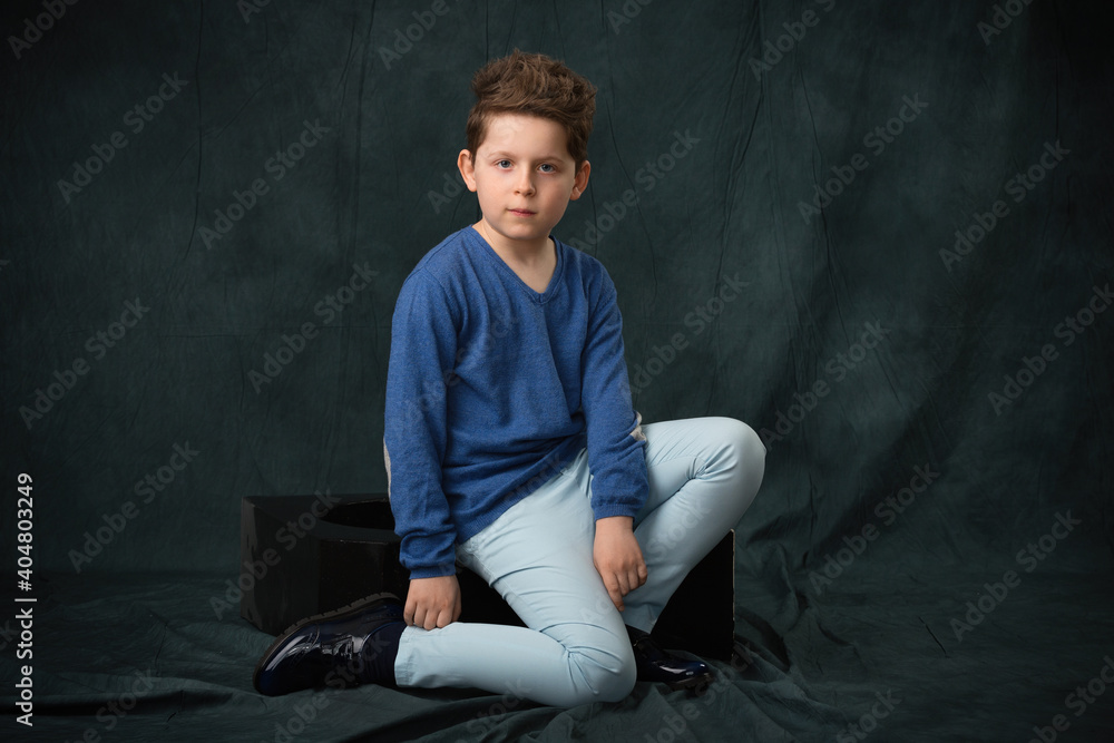 Obraz premium teen boy in a blue sweater and blue trousers posing while sitting in the studio on a gray fabric background