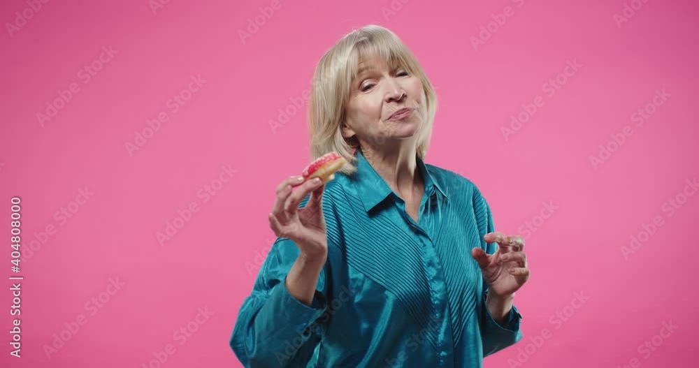 Portrait of cheerful pretty Caucasian senior blonde lady in blue shirt holding in hand donut eating and enjoying tasty delicious food standing on pink wall in studio with funny face expression. Portrait of cheerful pretty Caucasian senior blonde lady in blue shirt holding in hand donut eating and enjoying tasty delicious food standing on pink wall in studio with funny face expression.