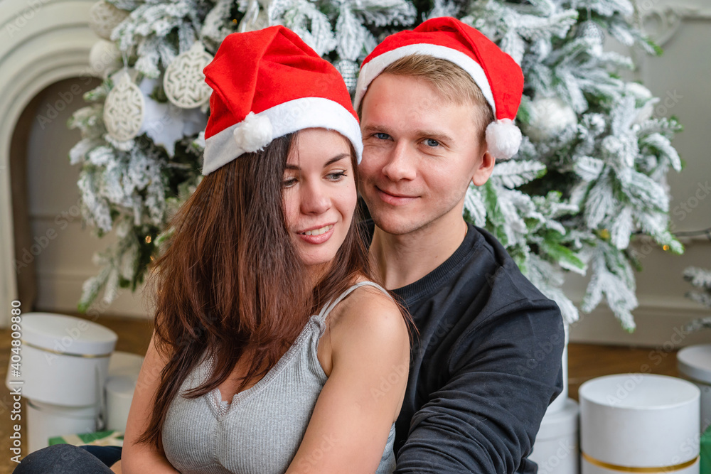 A loving couple of man and woman embrace next to a Christmas tree, dressed in Santa hats. A loving couple of man and woman embrace next to a Christmas tree, dressed in Santa hats.