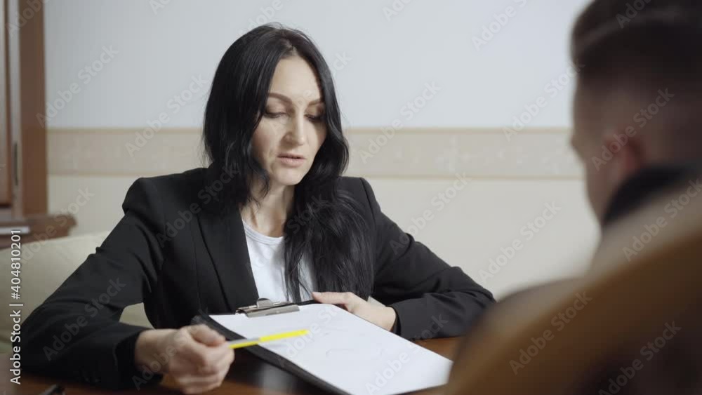Female Caucasian psychologist showing sad face on paper and talking to blurred man in office. Portrait of confident professional woman consulting patient with nervous breakdown. Female Caucasian psychologist showing sad face on paper and talking to blurred man in office. Portrait of confident professional woman consulting patient with nervous breakdown.