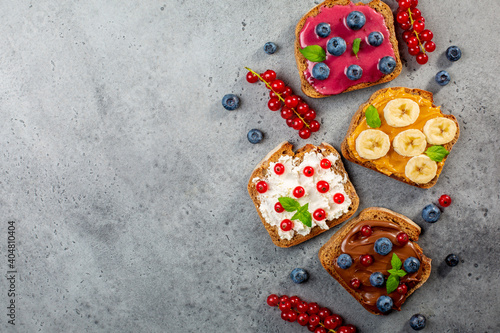 Variety of sweet sandwiches with peanut butter, chocolate butter, berry could, cream cheese, decorated with bananas, berries, mint. Grey background. Copy space.