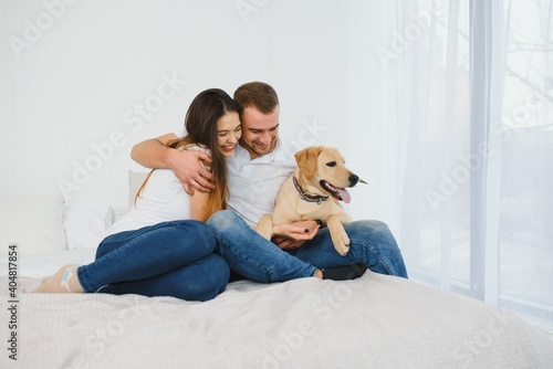 Cheerful brunette young female plays with her favourite dog in bed while her husband surfes social networks on laptop computer  spend weekends at home in bedroom. Domestic atmosphere