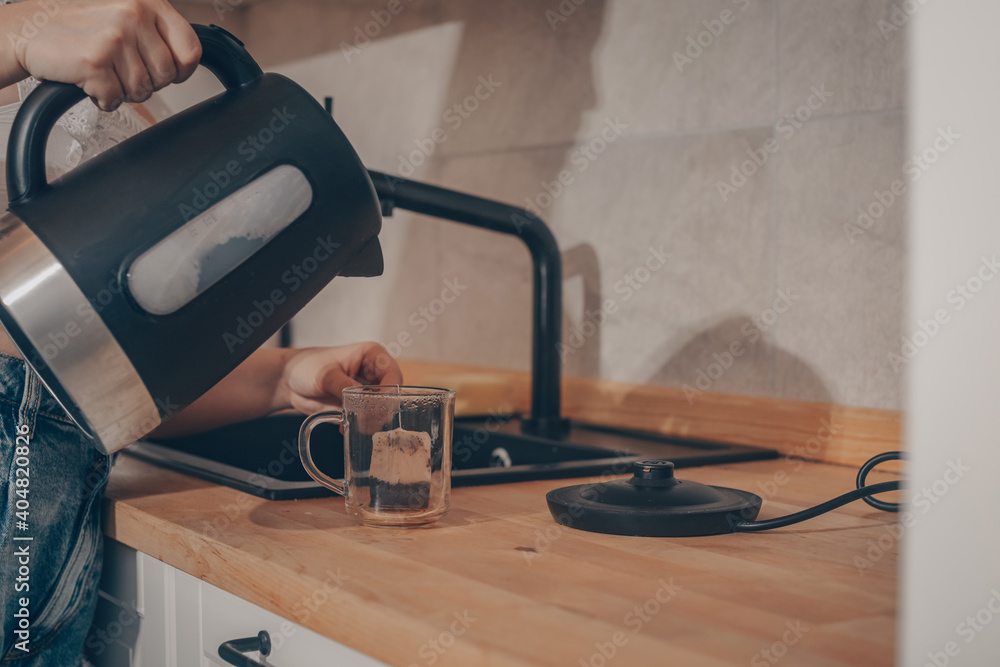 A black kettle is on the kitchen table. A woman's hand holds a teapot and makes tea. Tea bag in a mug with water, tea leaves. A black kettle is on the kitchen table. A woman's hand holds a teapot and makes tea. Tea bag in a mug with water, tea leaves.